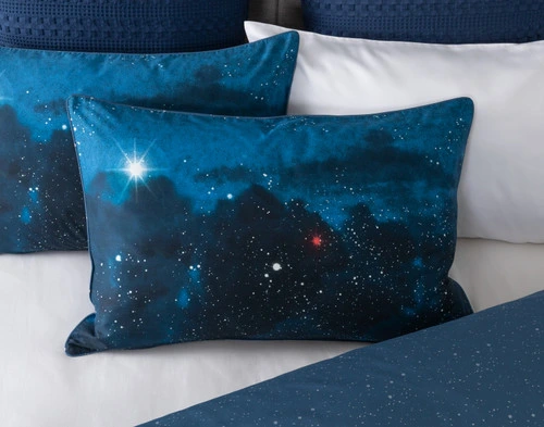 Vue de face de notre couvre-oreiller Supernova posé sur un couvre-oreiller assorti et sur le site blanc draps pour montrer son motif de ciel étoilé.