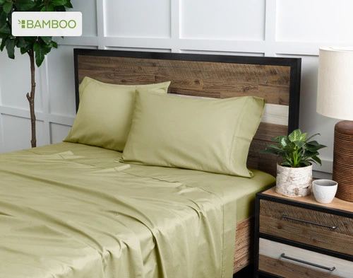 Vue en angle de notre Bamboo Cotton drap  ensemble  en Elm Green habillé au-dessus d'un lit en bois dans une chambre blanche remplie de plantes.