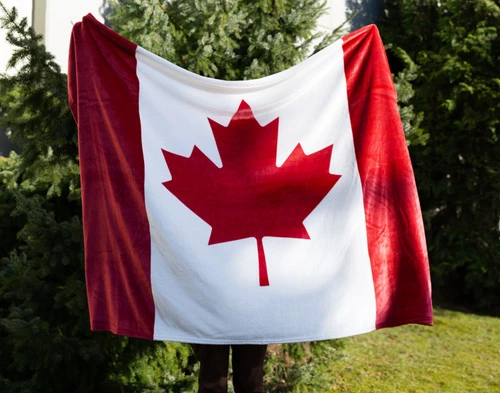 Front view of a person holding up our Canada Flag Fleece Throw in a green tree-filled space to show its traditional design.