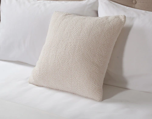 Angled view of our Ripple Boucle Square Cushion in Cream leaning against two white pillows on an undressed bed.