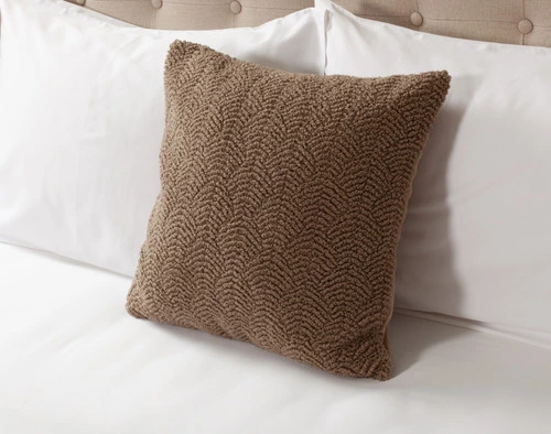 Angled view of our Ripple Boucle Square Cushion in Truffle leaning against two white pillows on an undressed bed.