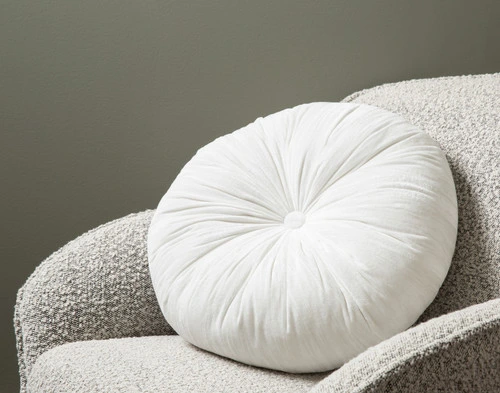 Angled view of our White Pleated Velvet Round Cushion leaning against the arm of a small grey armchair.