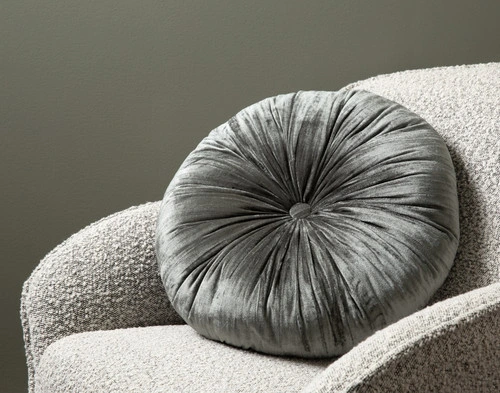 Angled view of our Fir Pleated Velvet Round Cushion leaning against the arm of a small grey armchair.