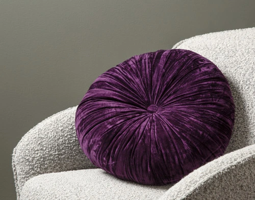 Angled view of our Purple Pleated Velvet Round Cushion leaning against the arm of a small grey armchair.