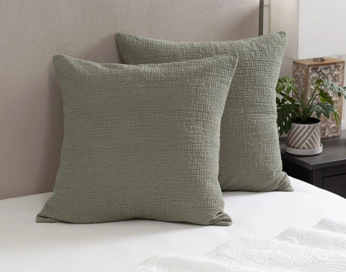 Angled view of our Crinkle Euro Shams in Sage leaning against a headboard on a bed dressed in white sheets.