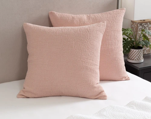 Angled view of our Crinkle Euro Shams in Peony leaning against a headboard on a bed dressed in white sheets.