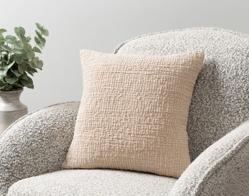 Our Crinkle Square Cushion Cover in Sand sitting on an armchair in a simple room.