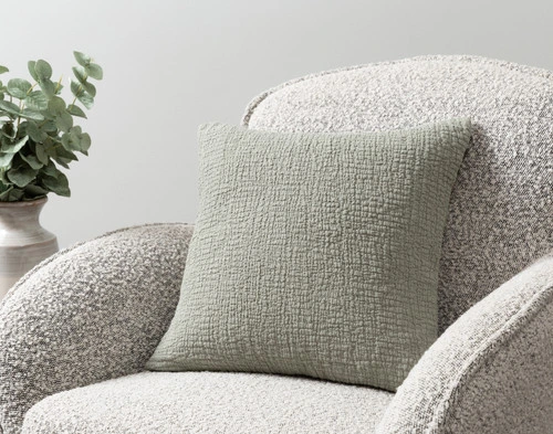 Our Crinkle Square Cushion Cover in Sage sitting on an armchair in a simple room.