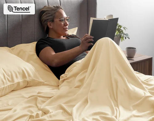 Woman sitting upright in a bed reading her book curled up in the flat sheet of our BeechBliss TENCEL™ Modal Sheet Set in Cornsilk.