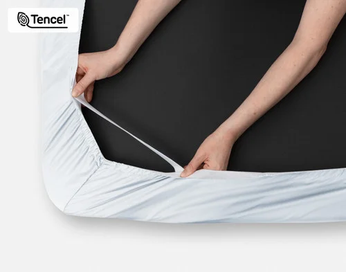 Close-up on two hands stretching the elastic snug fit band on the corner of our Eucalyptus Luxe TENCEL™ Lyocell Fitted Sheet in Blue Chalk.