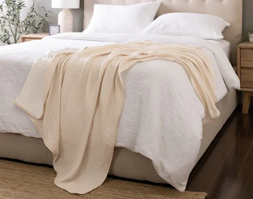 Our Bamboo Gauze Throw in Bone draped over the corner of a bed.