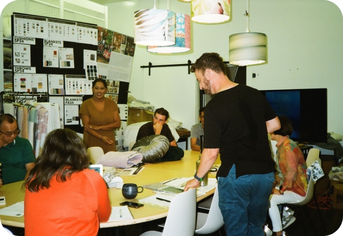 The QE Home product development team collaborates around a table, reviewing fabric samples and ideas. The creative studio features mood boards, textile samples, and a relaxed, focused atmosphere.