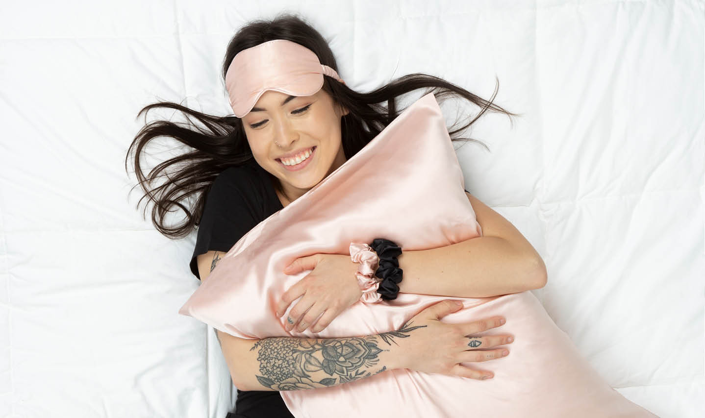 Top view of a cheerful woman with tattoos hugging a blush pink silk pillowcase. Top view of a cheerful woman with tattoos hugging a blush pink silk pillowcase.
