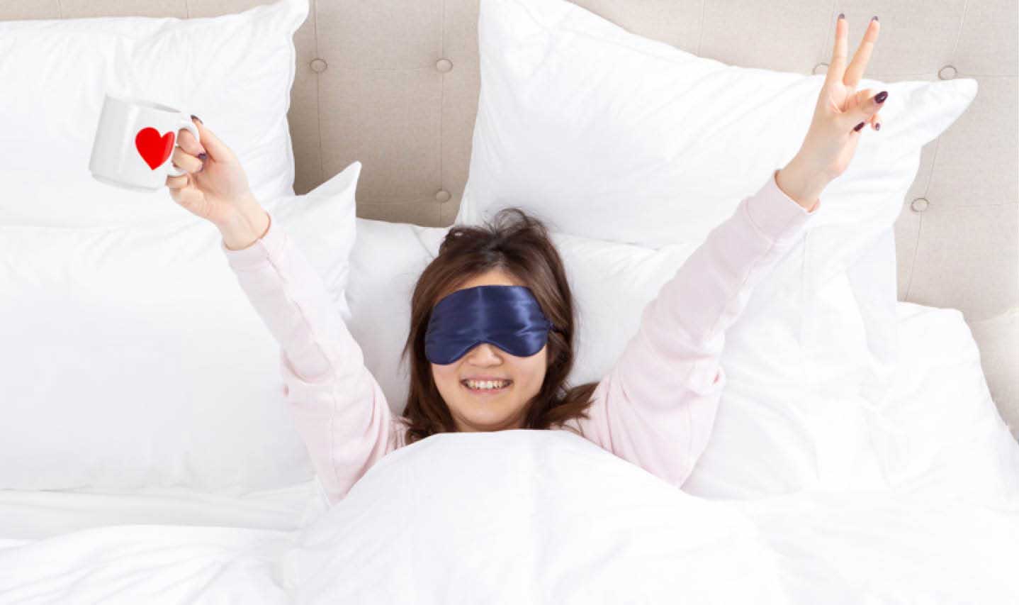 Front view of a cheerful woman with her arms in the air while resting in a white bed, wearing a silk eye mask. Front view of a cheerful woman with her arms in the air while resting in a white bed, wearing a silk eye mask.