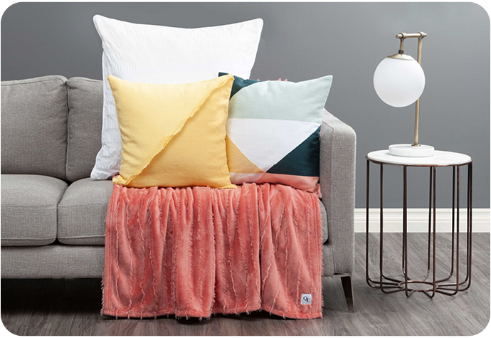 An assortment of coloured cushions and an orange velvet throw are placed on a grey sofa next to a small table and lamp