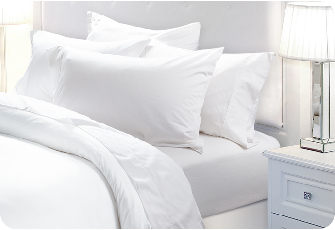Bed with white sheet set and four pillows next to a bedside table and lamp