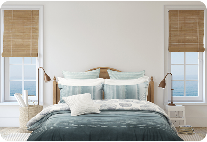 Blue bedding set on a styled bed with two lamps and two windows dressed with blinds showing an ocean in the distance