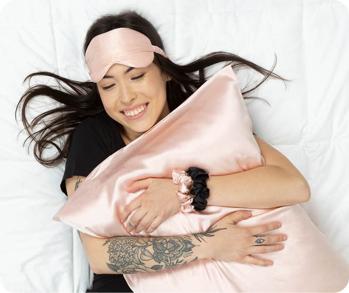 A person lies down hugging one of our Mulberry Silk Pillowcases in Blush, wearing an eye mask in the same fabric and colour.s