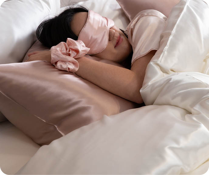 A person rests in bed surrounded with our Mulberry Silk bedding, donning our Silk Moon accessories.