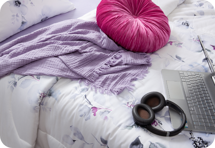 A bed with our floral Talia Comforter Set, our Muslin Gauze Throw in Lilac, our pink Sulani Round Cushion, a laptop, and headphones.