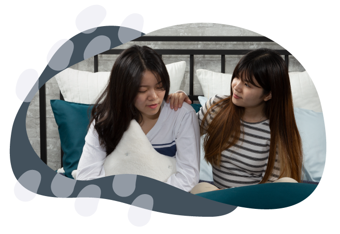 Woman holding a cushion sitting in bed, leaning on and being comforted by a friend beside her.