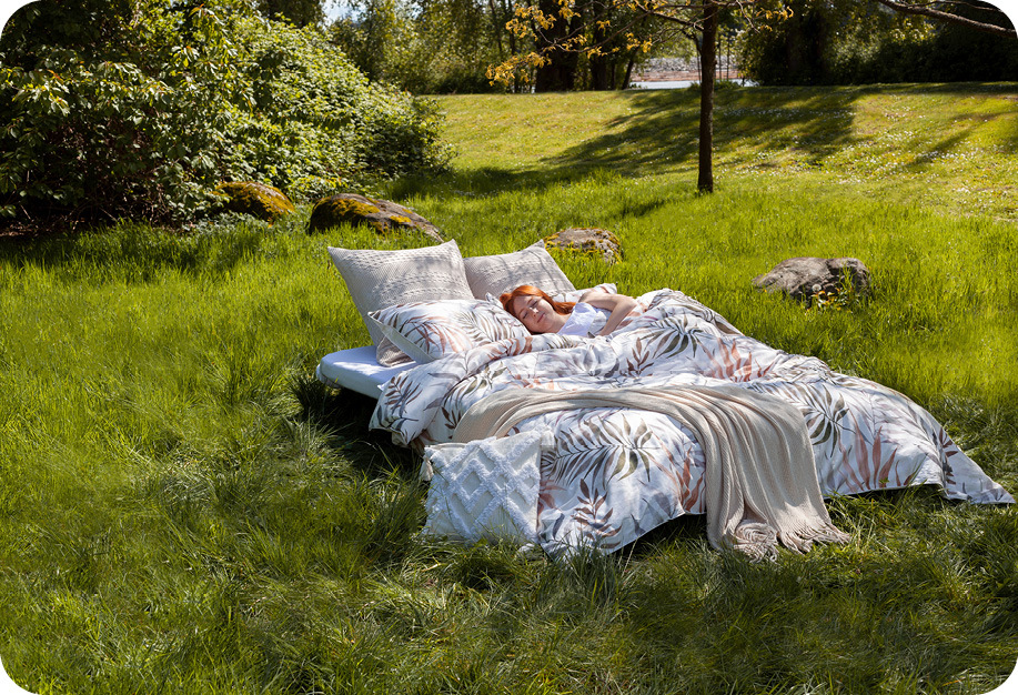 Une personne dormant dans un lit habillé de notre collection San Luca, de la housse de coussin carrée à losanges et de la chenille Jeté en naturel, entourée d'une prairie. 