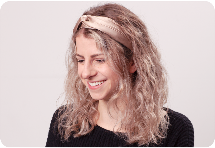 Blonde woman wearing a Silk Twist Headband with bronze colour while smiling downward.
