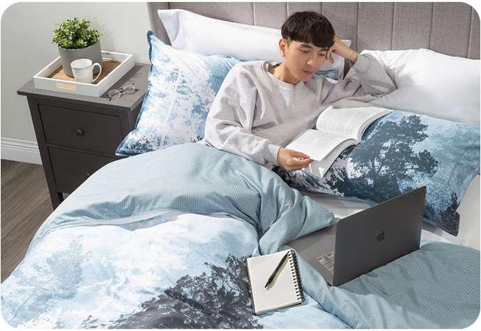 Un homme est allongé dans un lit équipé de notre housse de couette Cypress et de nos taies d'oreiller. Il est allongé et lit un livre avec un ordinateur portable et un journal à côté de lui.