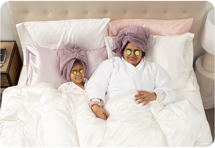 Une mère et sa fille sont représentées allongées dans leur lit, des tranches de concombre couvrant leurs yeux et leurs cheveux étant enveloppés dans des serviettes. Nos taies d'oreiller Soie de mûrier sont représentées sur le lit blanc.