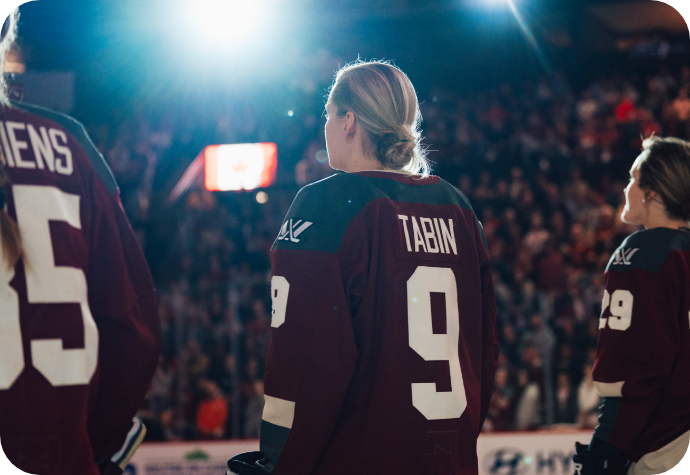 Hockey player Kati Tabin wearing the official PWHL Montreal maroon jersey with the name "Tabin" facing away from the camera, with teammates. Bright arena lights shine above, and a blurred crowd is visible in the background.