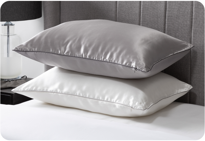 Angled view of two pillows in white and silver mulberry silk pillow shams, stacked on top of each other on an empty white bed.