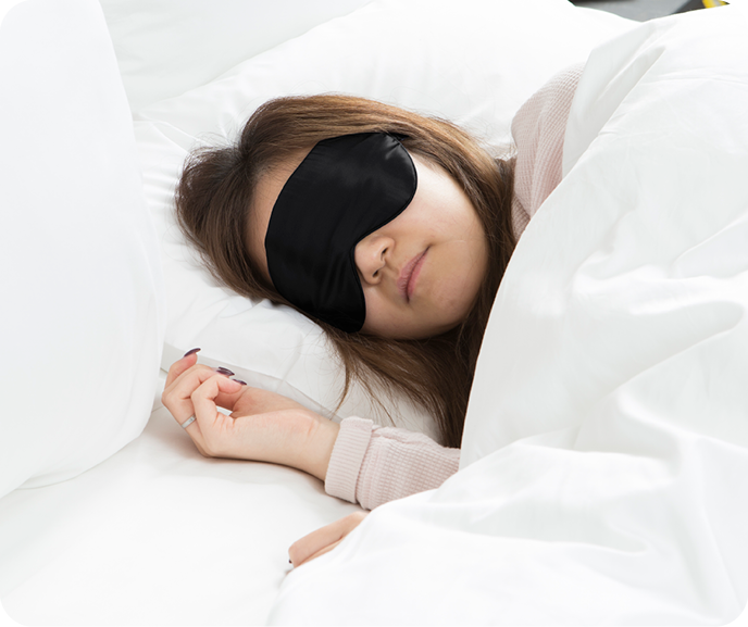 A person wearing a black sleep mask is lying in bed, partially covered by a white duvet.